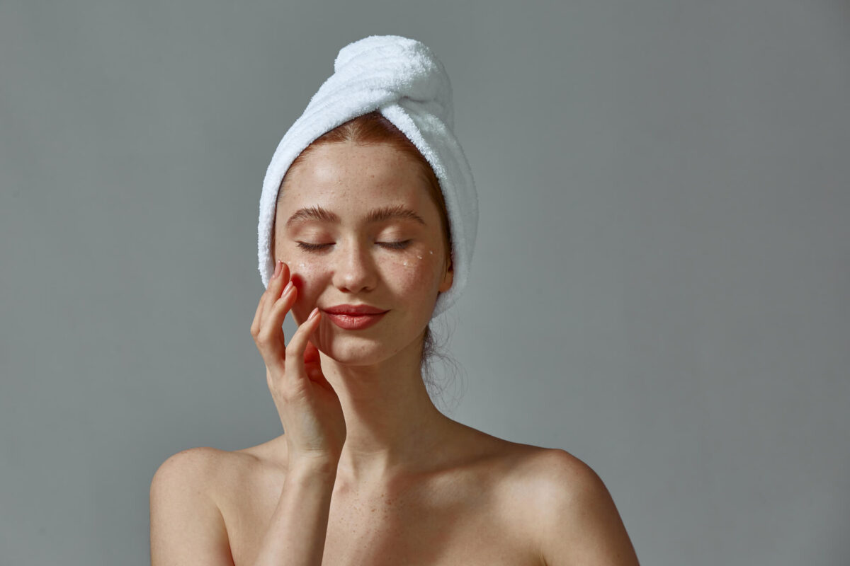 Happy girl in towel touch perfect face skin applying nourishing cream